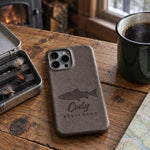 Country Gray Cody STEELHEAD iPhone case in full-grain leather on a knotty pine cabin table beside an open fly box and a green enamel coffee mug
