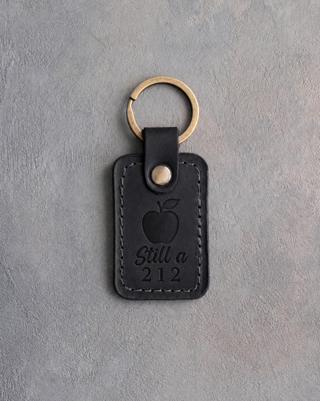 Black leather keychain debossed with a solid apple above Still a and 212, on gray concrete