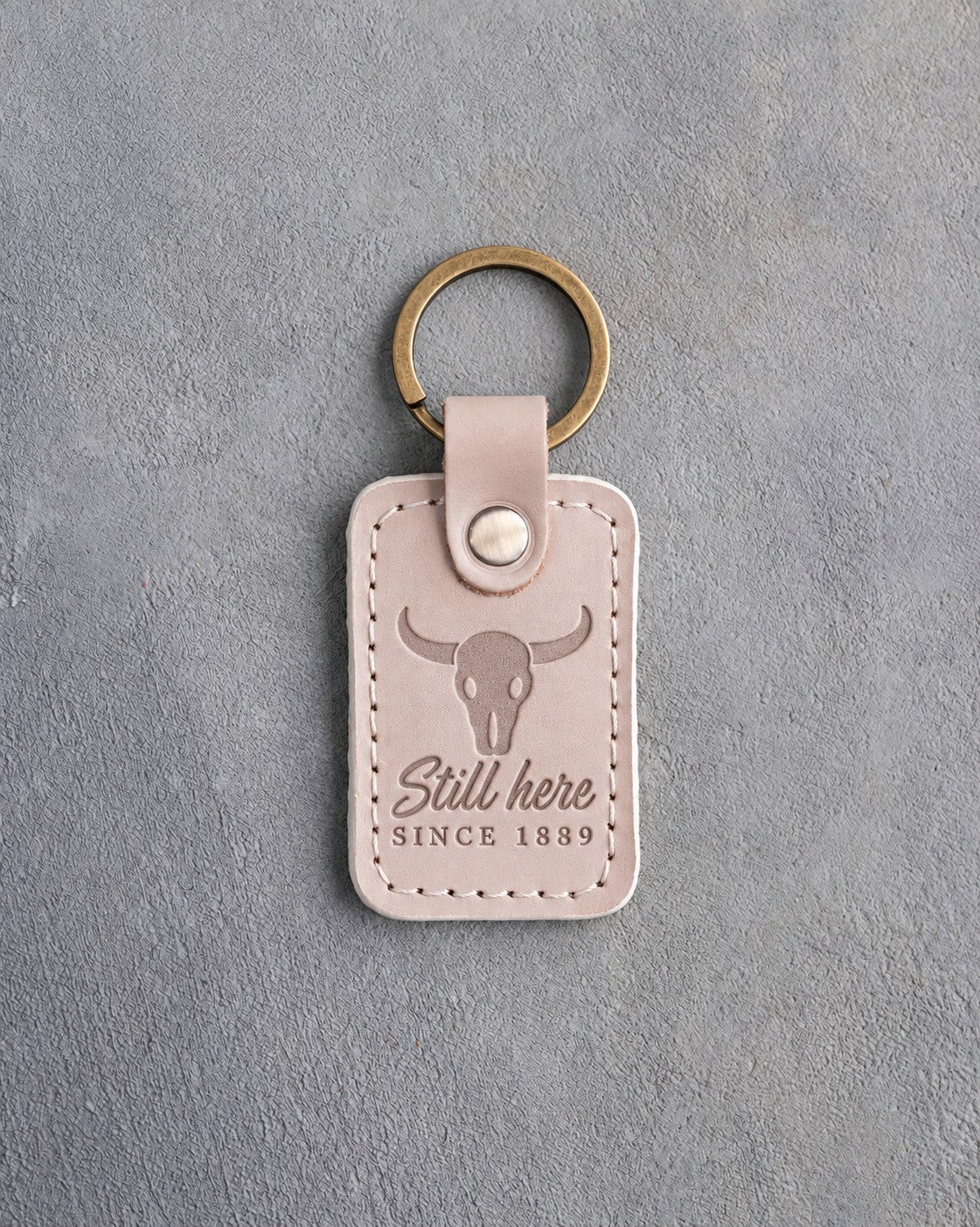 Gray sand leather keychain debossed with a steer skull above Still here and SINCE 1889, on gray plaster