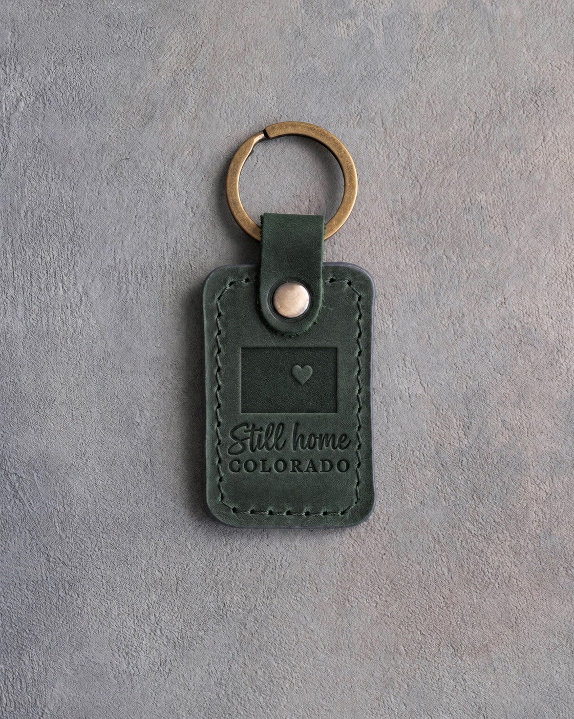 Forest green leather keychain debossed with a solid Colorado rectangle and a heart at Denver above Still home and COLORADO, on gray concrete