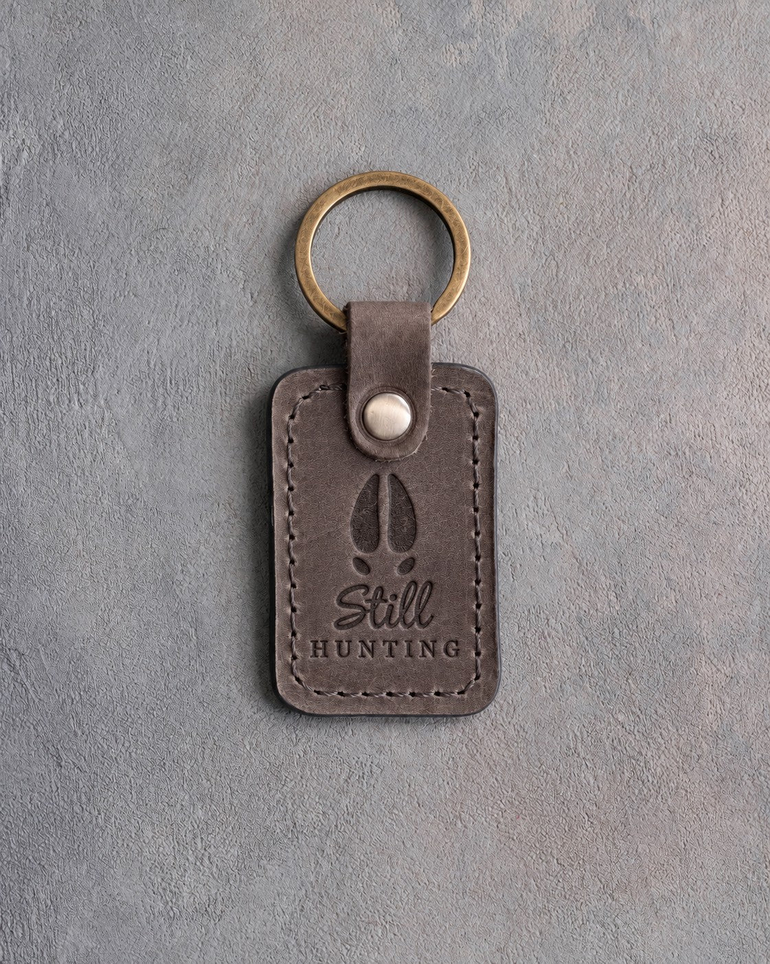 Country gray leather keychain debossed with a whitetail deer track above the words Still HUNTING, on gray plaster