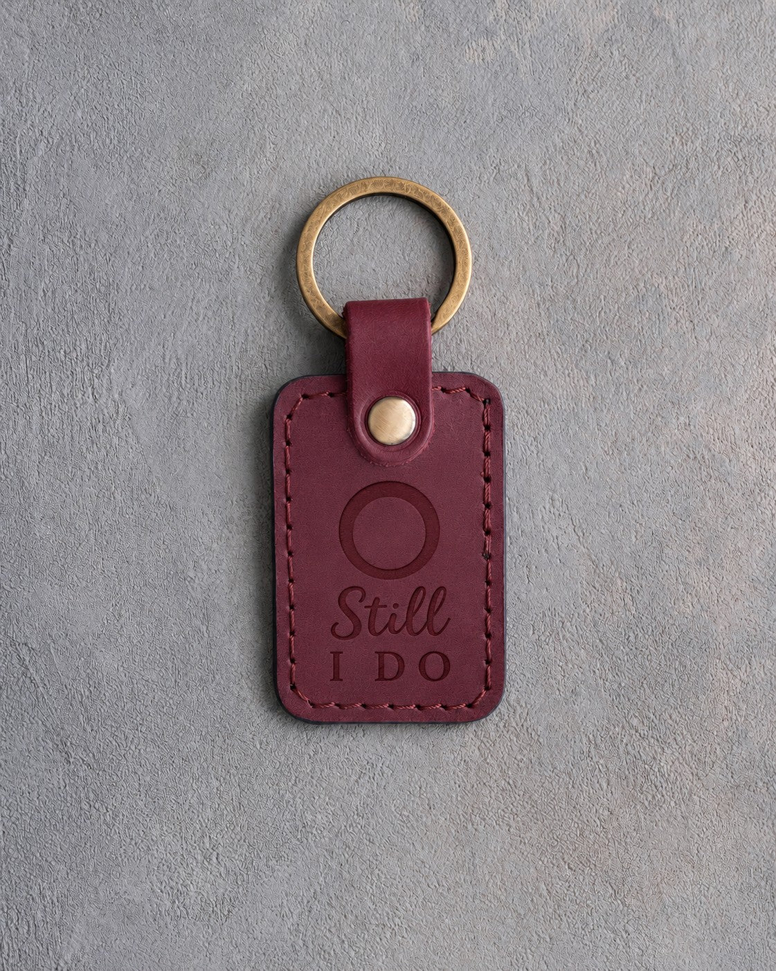 Sangria red leather keychain debossed with a single wedding band above the words Still I DO, on gray concrete