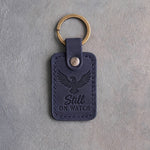 Blueberry navy leather keychain debossed with an eagle in flight above the words Still ON WATCH, on gray concrete
