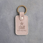 Gray sand leather keychain debossed with a solid five-point star above the words Still SHINING, on gray concrete