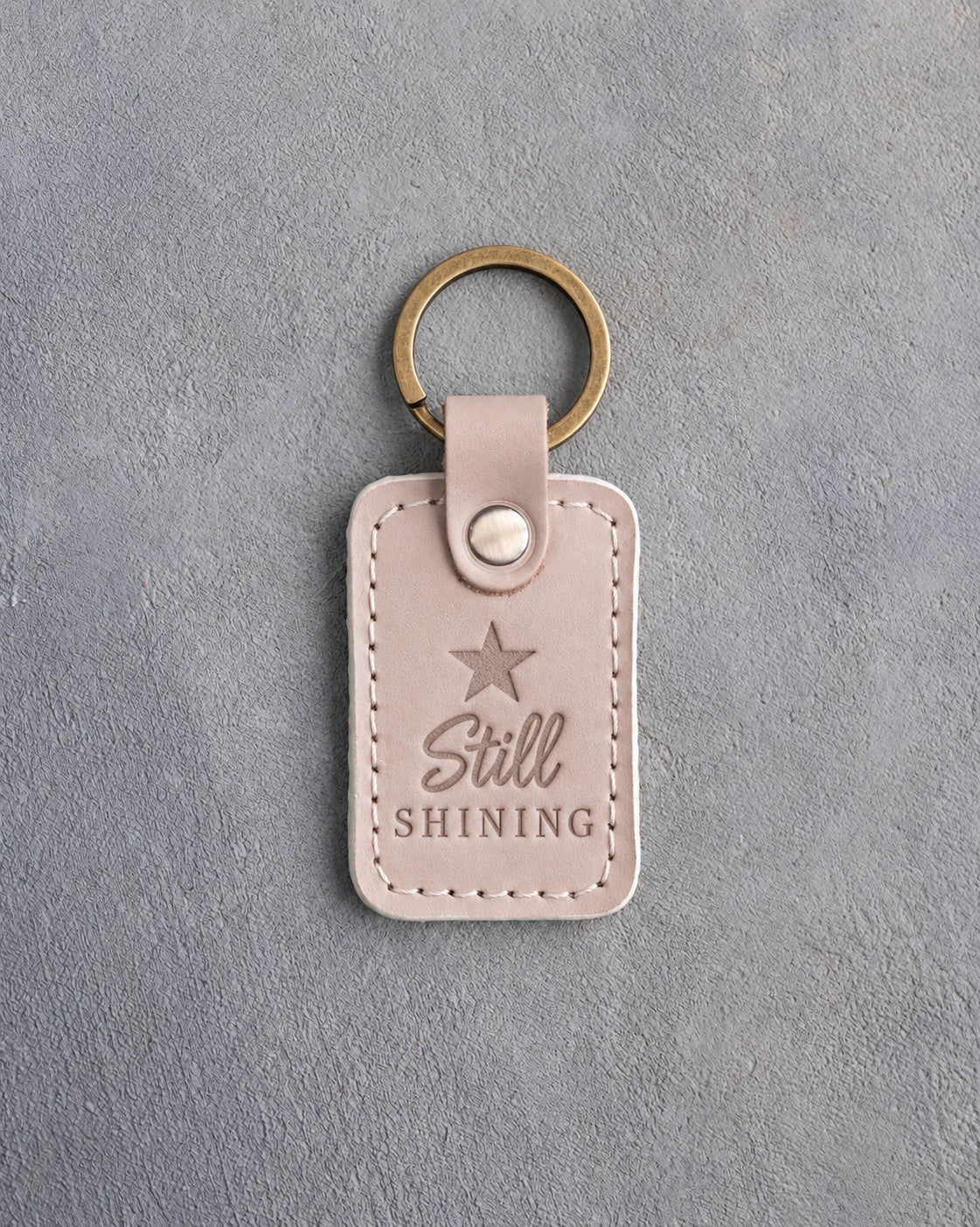 Gray sand leather keychain debossed with a solid five-point star above the words Still SHINING, on gray concrete