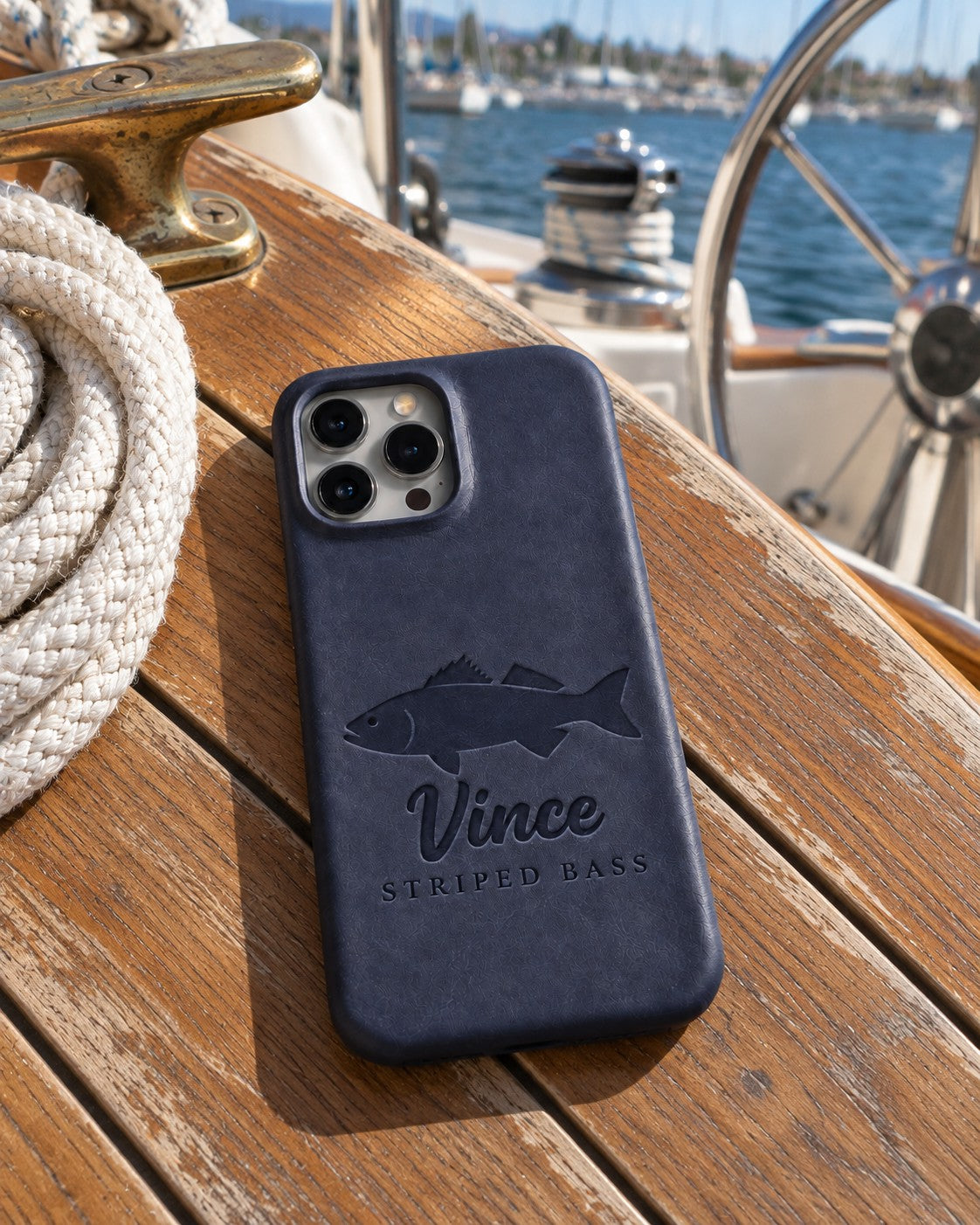 Blueberry Vince STRIPED BASS iPhone case in full-grain leather on the worn teak cockpit table of a sailboat beside a coiled white dock line