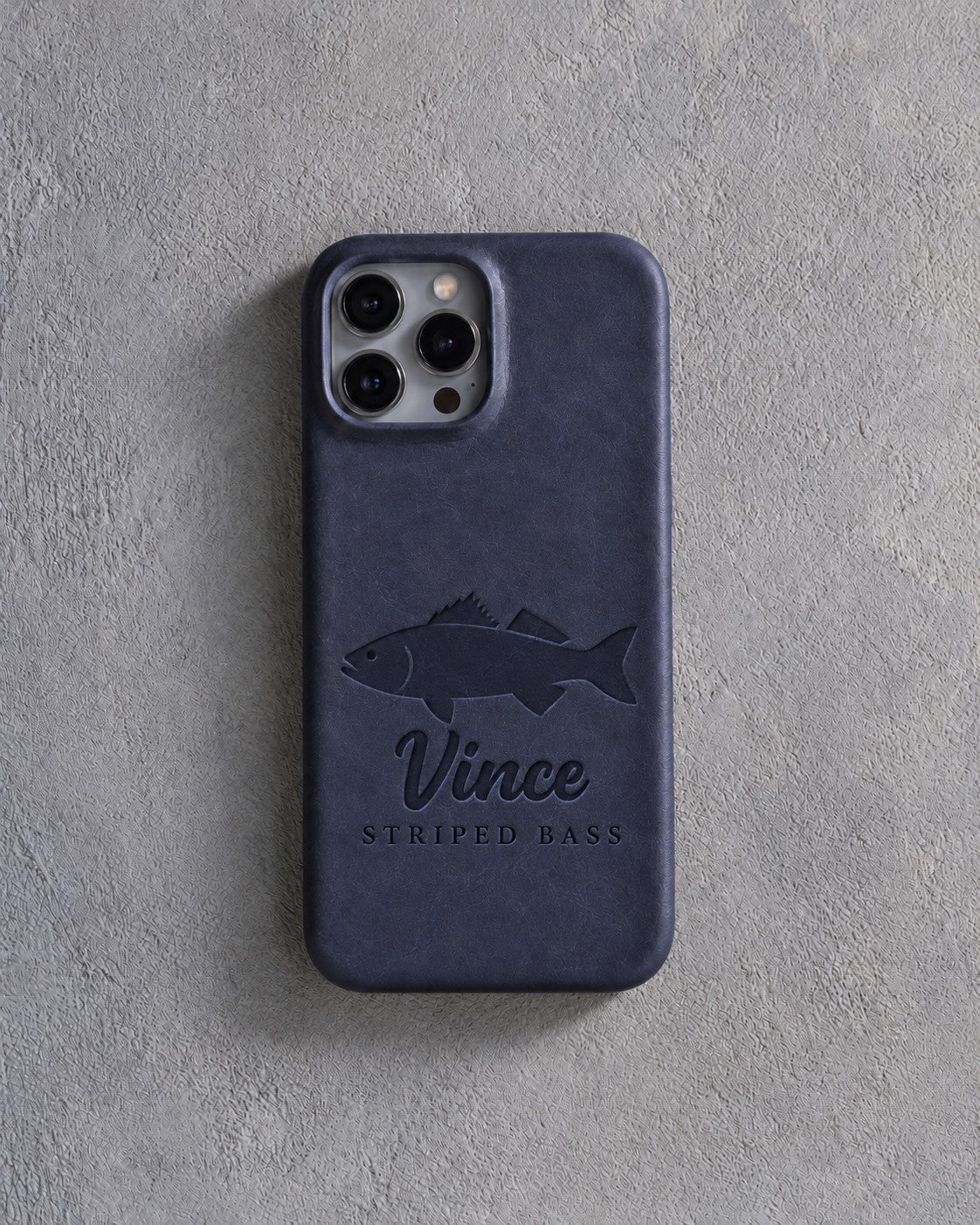 Back of a blueberry leather iPhone case debossed with a striped bass above Vince and STRIPED BASS below the camera, on gray plaster