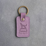 Lilac leather keychain debossed with a dog standing on a hilltop and the words Summit FOUR PAWS UP, on gray concrete