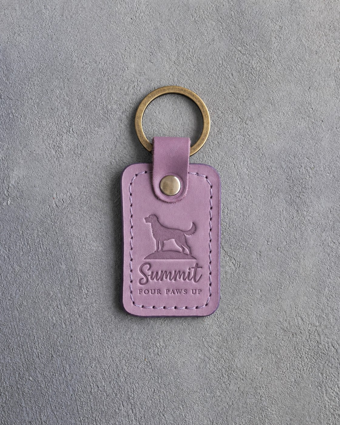 Lilac leather keychain debossed with a dog standing on a hilltop and the words Summit FOUR PAWS UP, on gray concrete