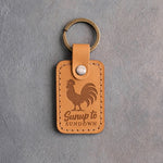 Indian Summer tan leather keychain debossed with a crowing rooster above the words Sunup to SUNDOWN, on gray concrete