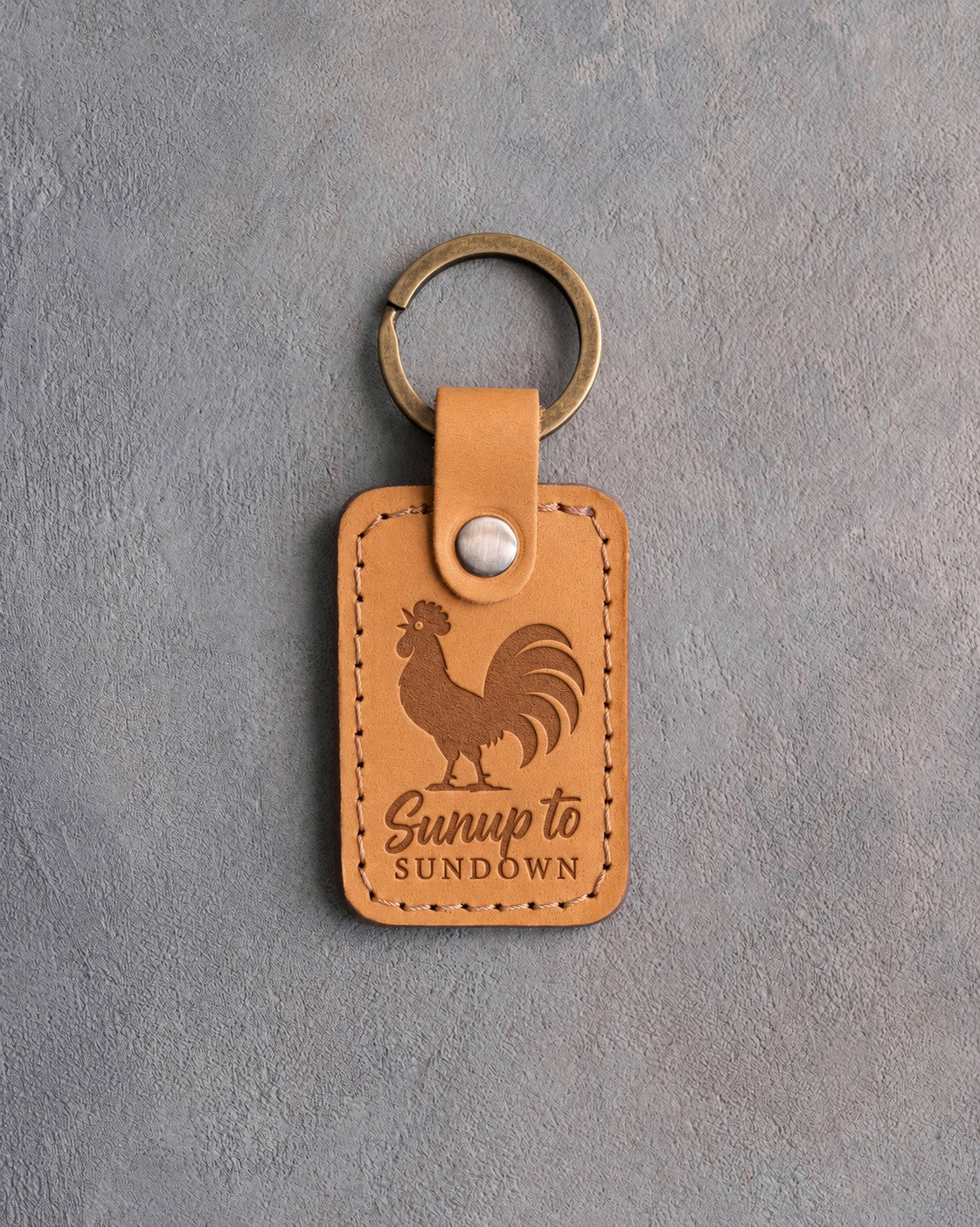 Indian Summer tan leather keychain debossed with a crowing rooster above the words Sunup to SUNDOWN, on gray concrete