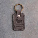 Country gray leather keychain debossed with a loafing cat above Life with a and TABBY CAT, on gray concrete