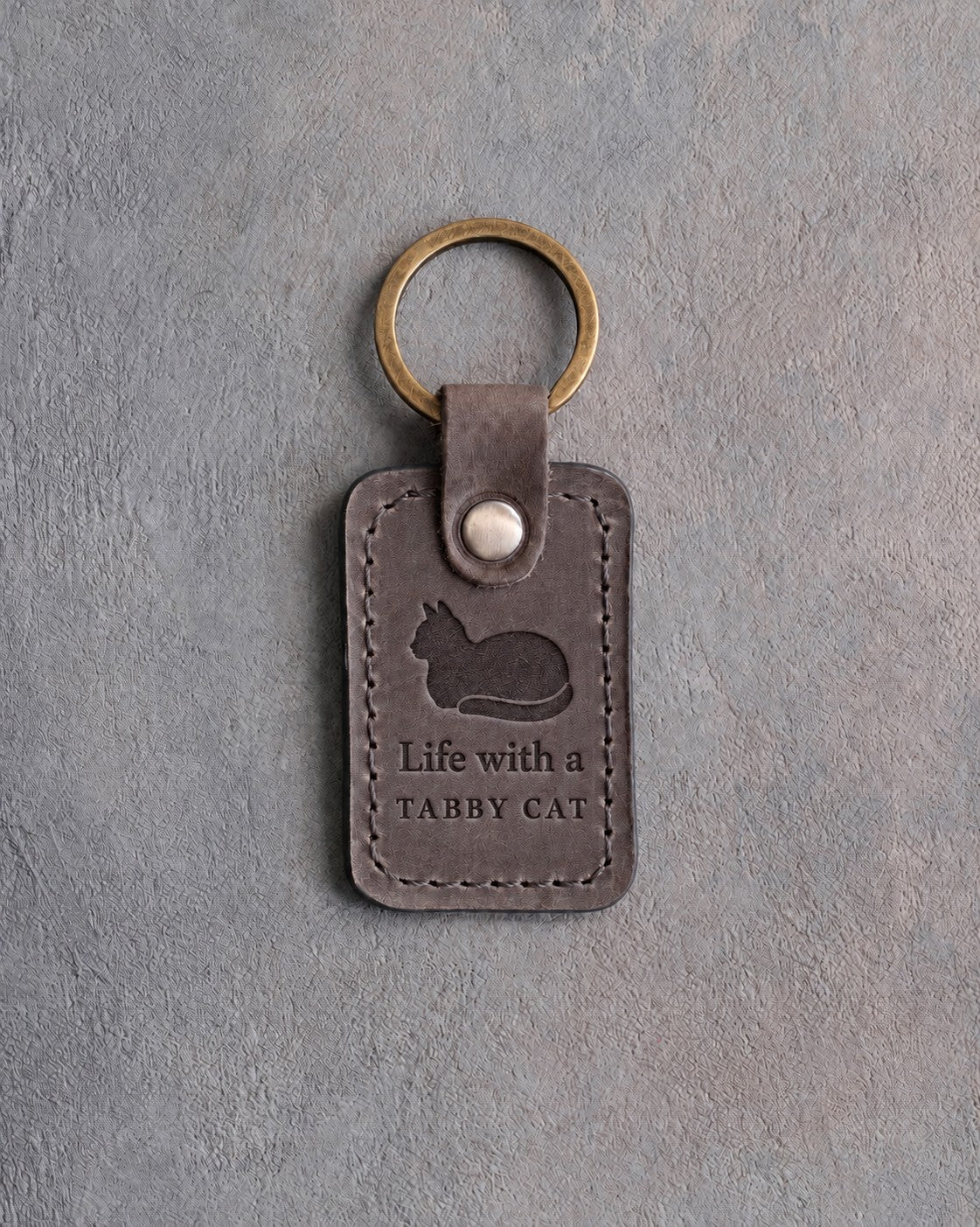 Country gray leather keychain debossed with a loafing cat above Life with a and TABBY CAT, on gray concrete
