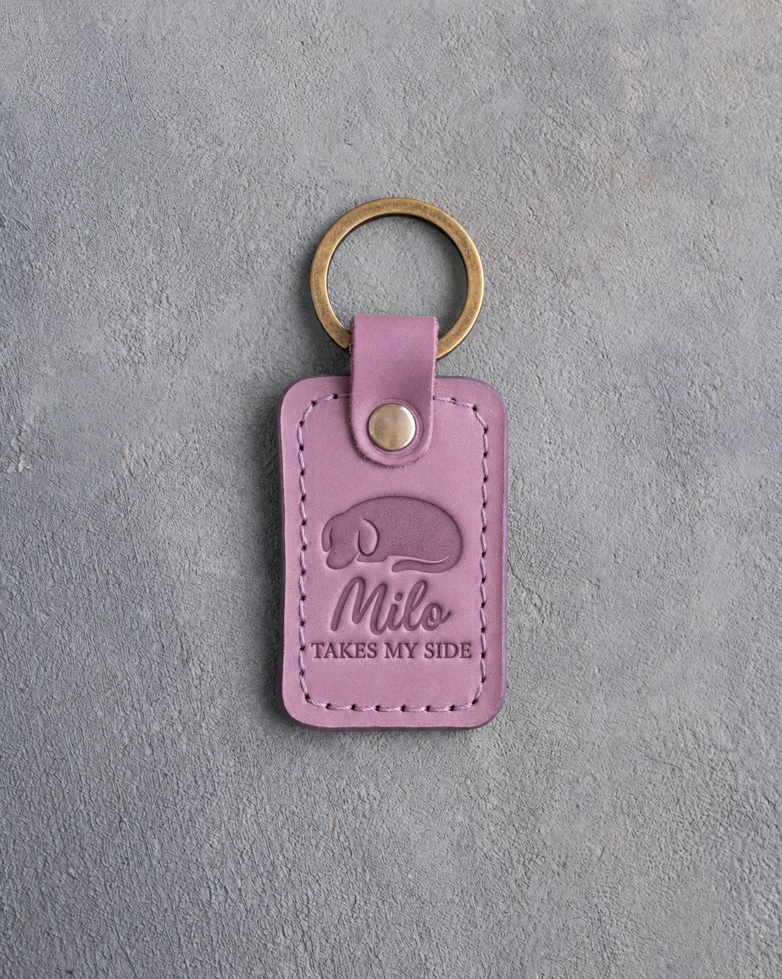 Lilac leather keychain debossed with a curled sleeping dog above Milo and TAKES MY SIDE, on gray concrete