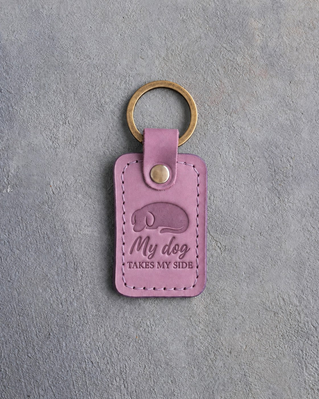 Lilac leather keychain debossed with a curled sleeping dog above My dog and TAKES MY SIDE, on gray concrete