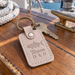 Gray sand Tarpon DAD keychain in full-grain leather on the worn teak cockpit table of a sailboat beside a coiled white dock line