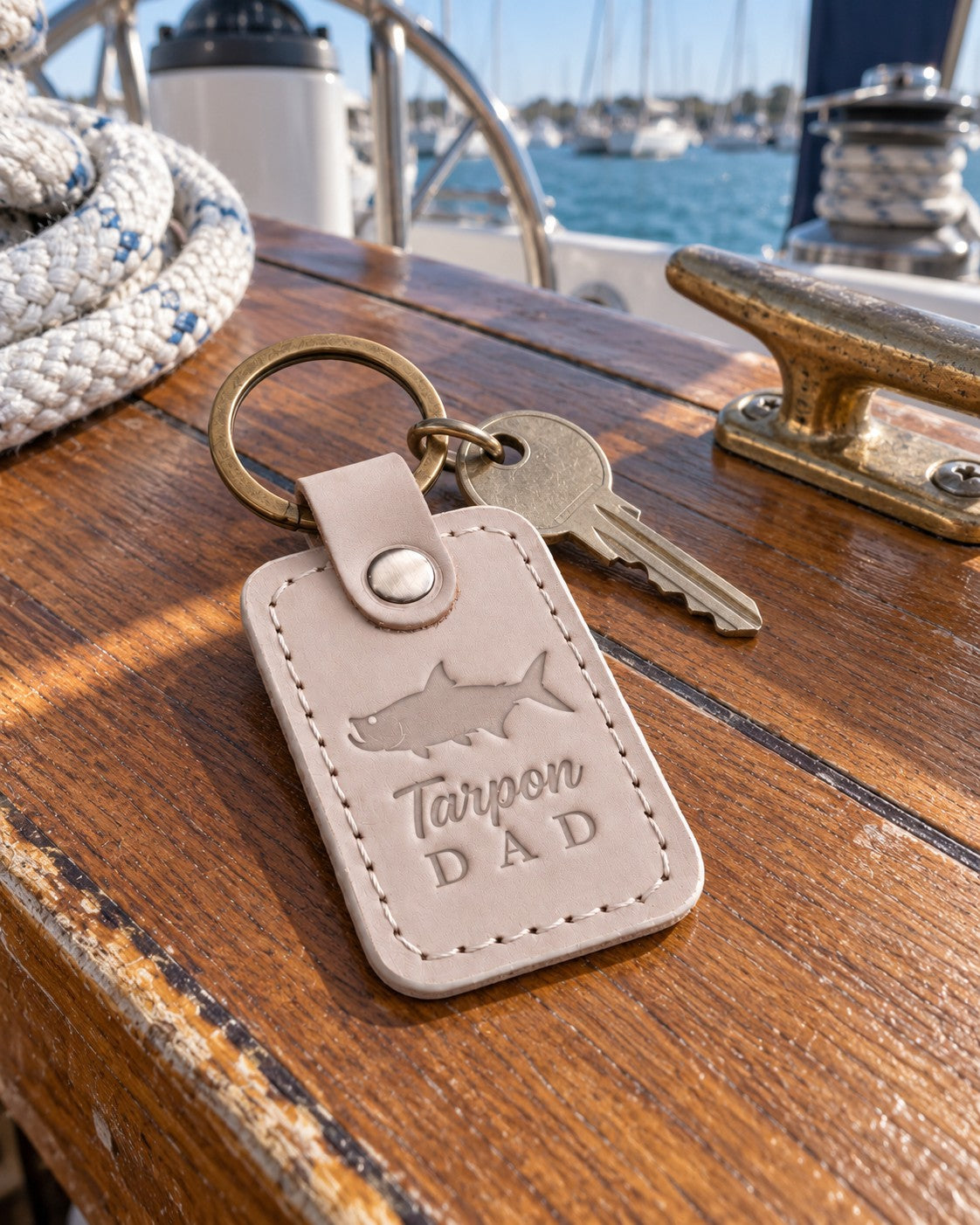 Gray sand Tarpon DAD keychain in full-grain leather on the worn teak cockpit table of a sailboat beside a coiled white dock line