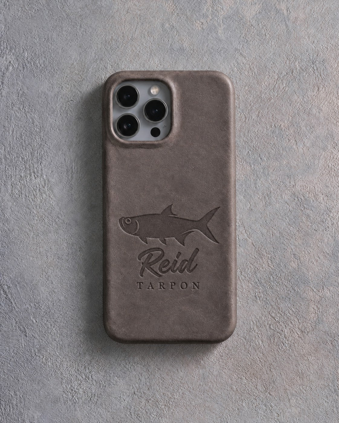 Back of a country gray leather iPhone case debossed with a tarpon above Reid and TARPON below the camera, on gray plaster