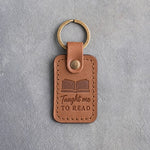 Caramel leather keychain debossed with an open book above Taught me and TO READ, on gray concrete