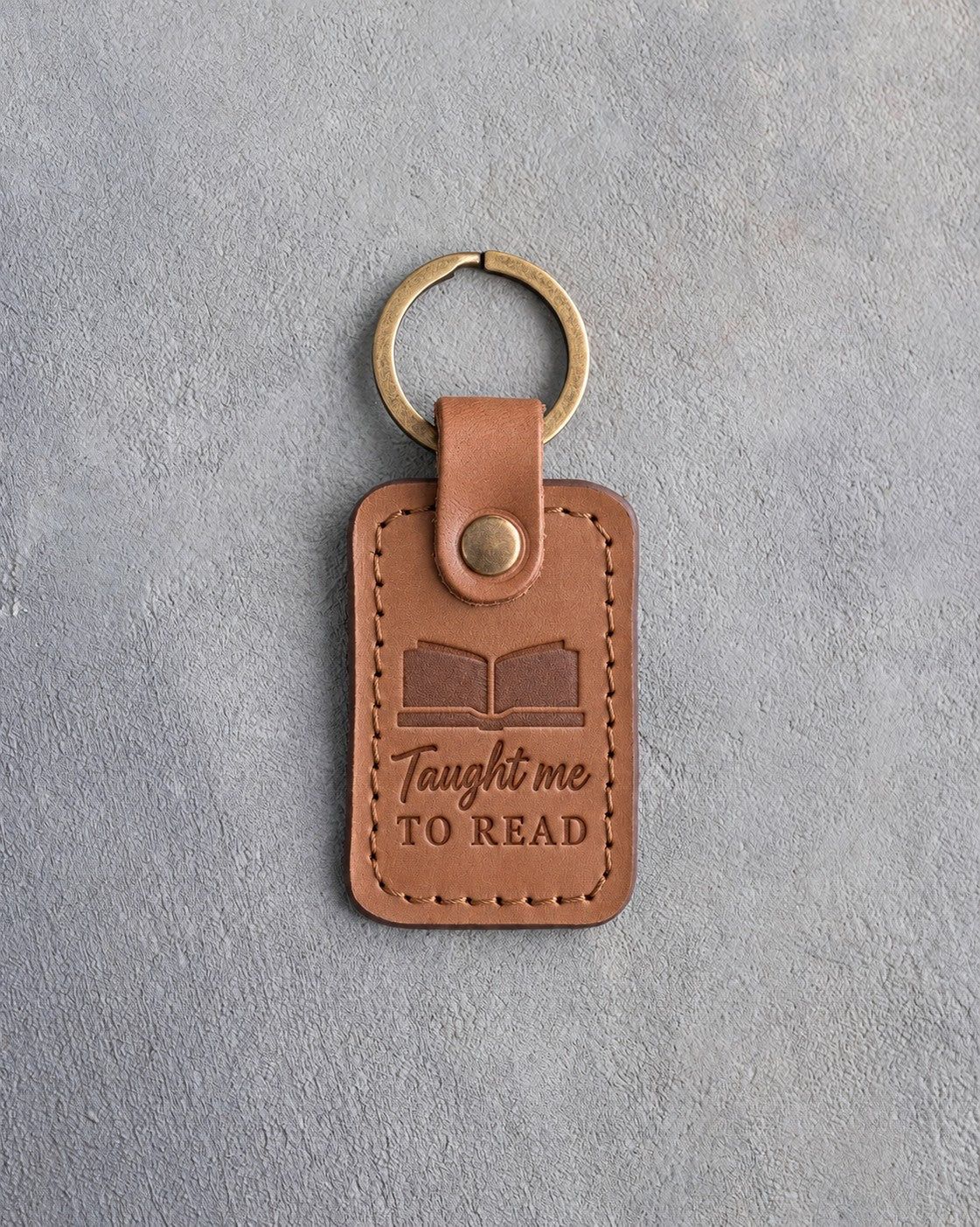 Caramel leather keychain debossed with an open book above Taught me and TO READ, on gray concrete