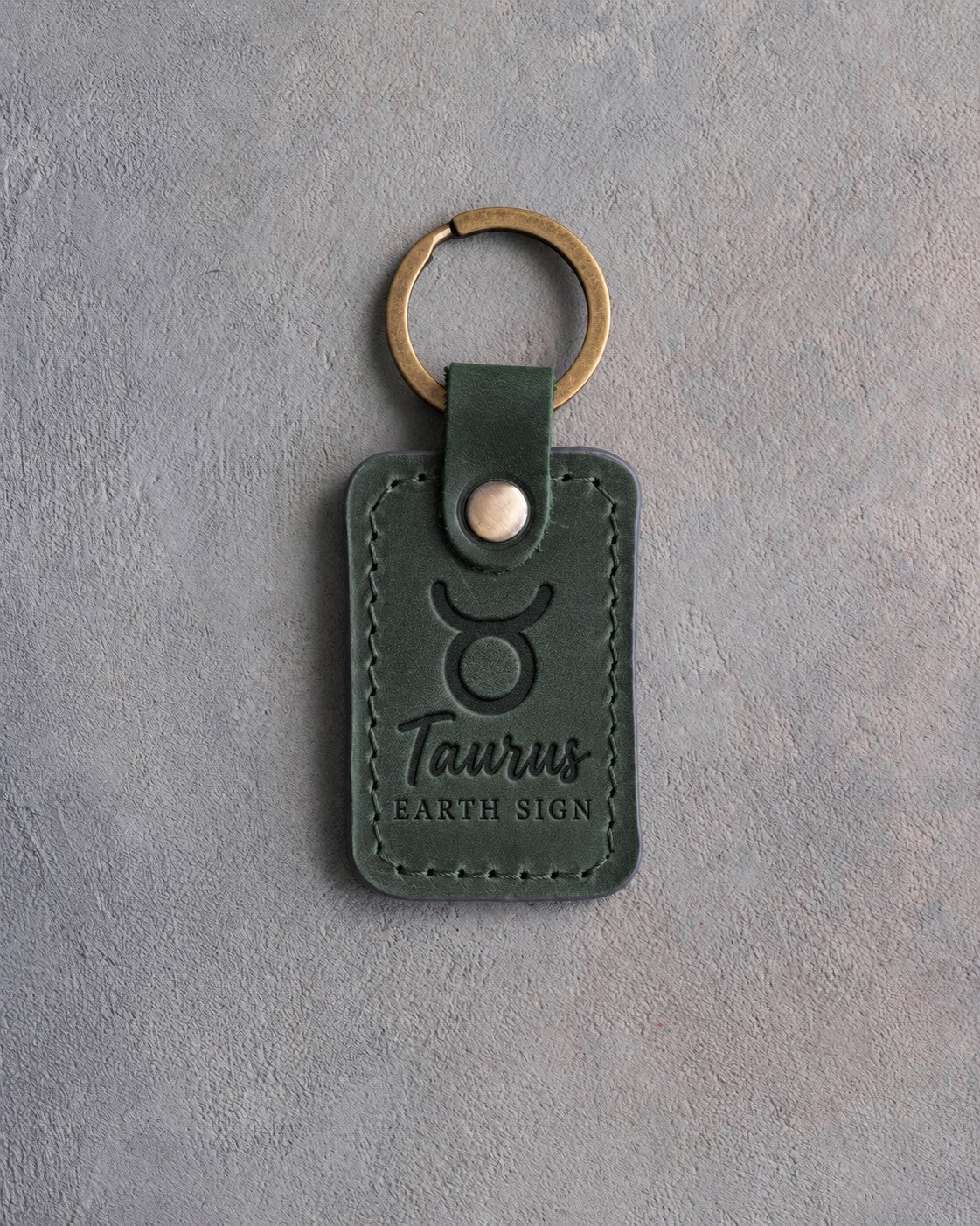 Forest green leather keychain debossed with the Taurus zodiac glyph above Taurus and EARTH SIGN, on gray concrete