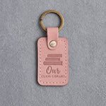 Blush pink leather keychain debossed with a stack of books above Our and CLASS LIBRARY, on gray concrete