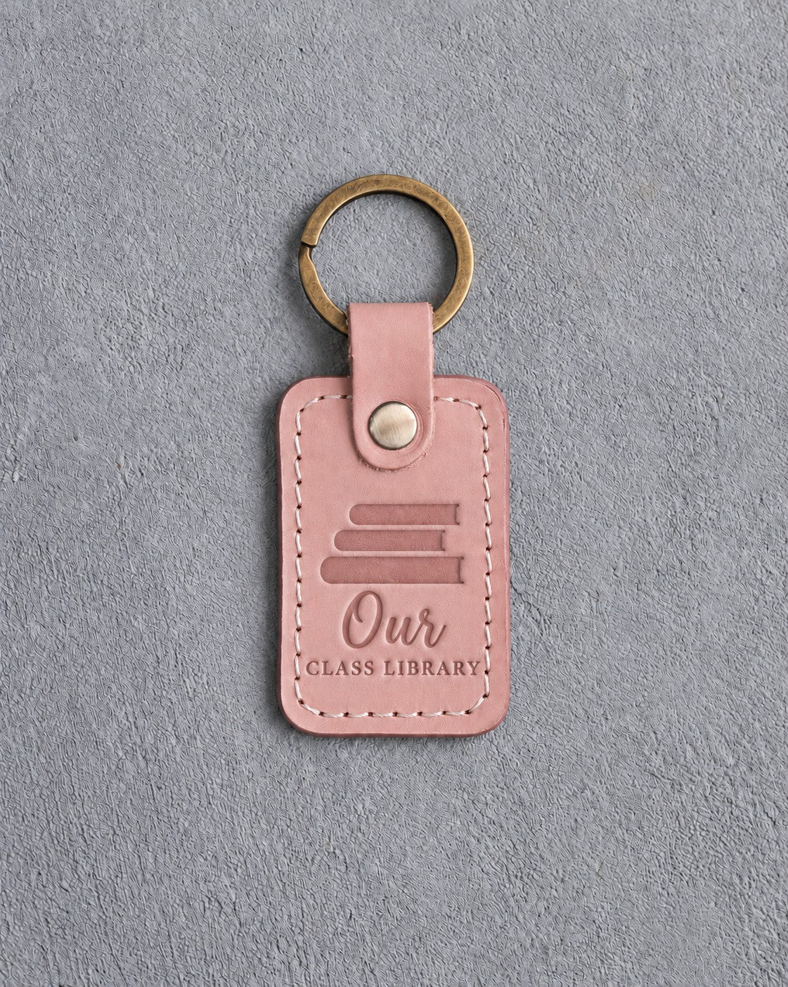Blush pink leather keychain debossed with a stack of books above Our and CLASS LIBRARY, on gray concrete