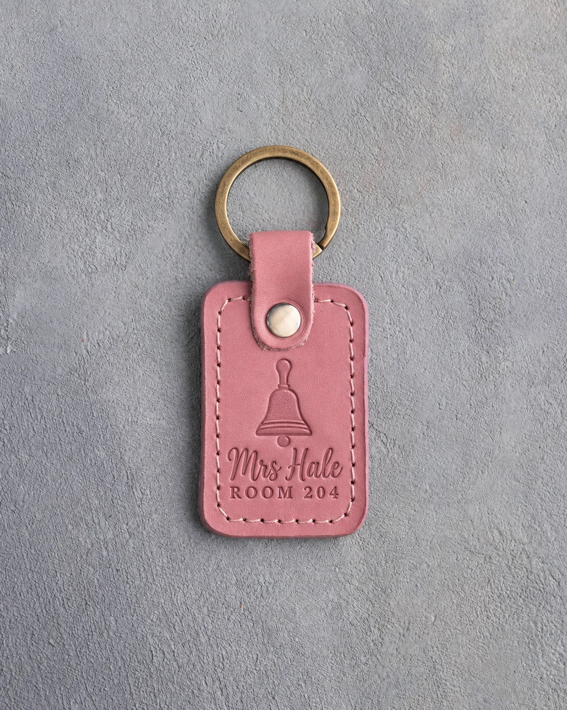 Rose pink leather keychain debossed with a hand bell above the name Mrs Hale and ROOM 204, on gray plaster
