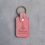 Rose pink leather keychain debossed with a hand bell above the words Thank you and TEACHER, on gray concrete