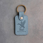 Turquoise leather keychain debossed with a duck cupped to land above the words Teal SEASON, on gray plaster