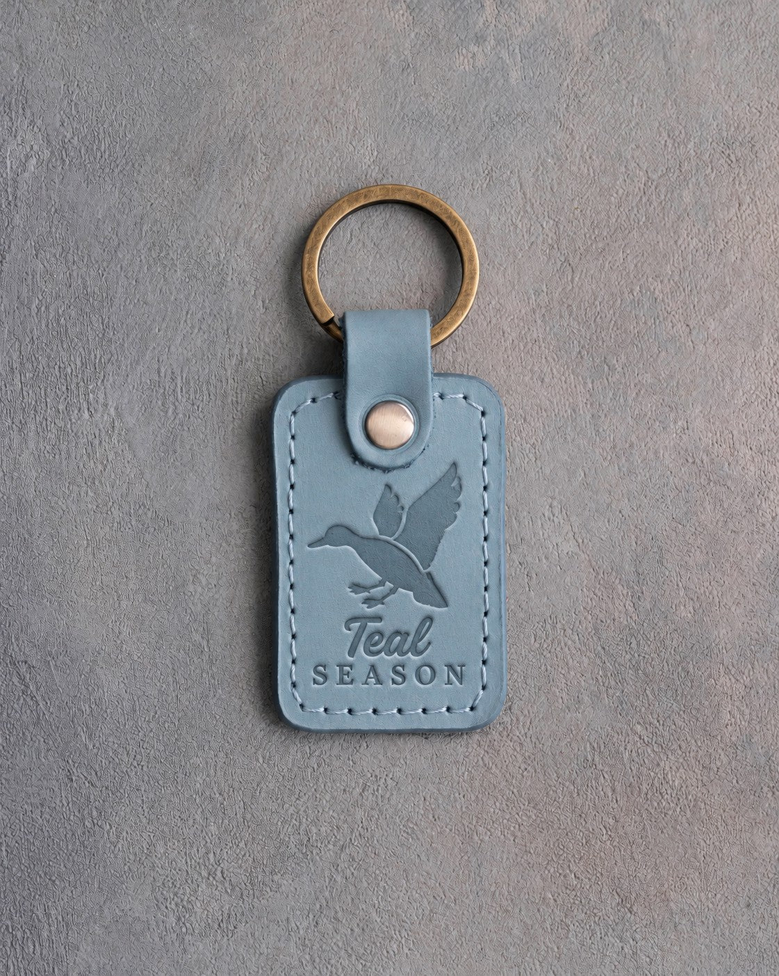 Turquoise leather keychain debossed with a duck cupped to land above the words Teal SEASON, on gray plaster
