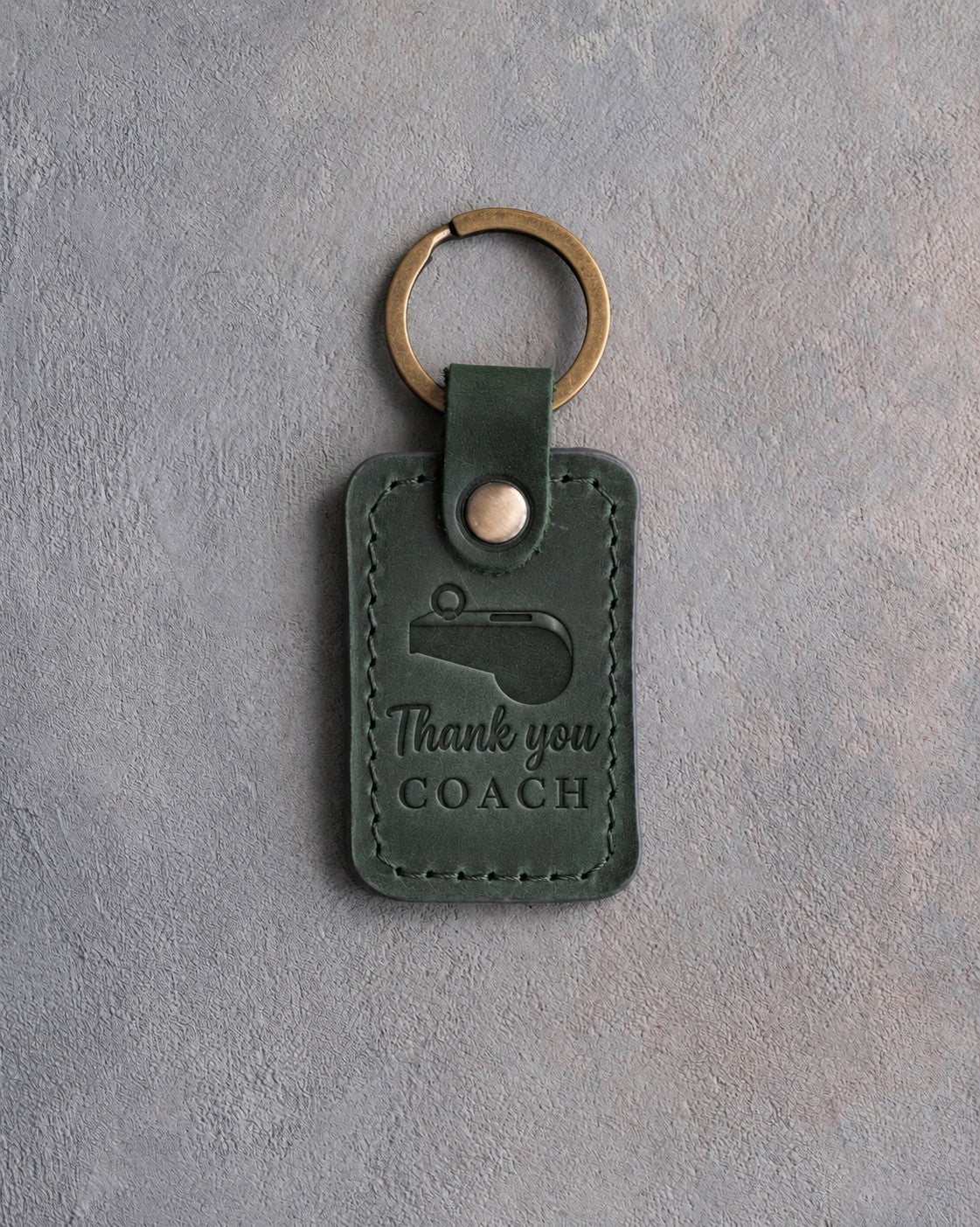 Forest green leather keychain debossed with a coach's whistle and the words Thank you COACH, on gray plaster