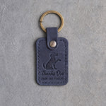 Blueberry navy leather keychain debossed with a dog raising its paw above Thanks Doc and FROM THE PATIENT, on gray concrete