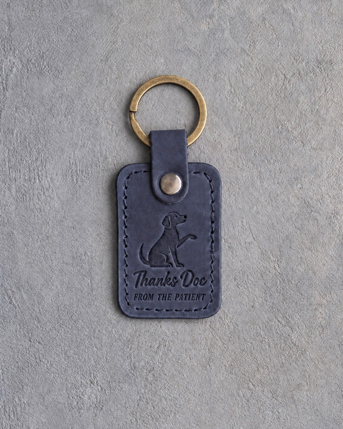 Blueberry navy leather keychain debossed with a dog raising its paw above Thanks Doc and FROM THE PATIENT, on gray concrete