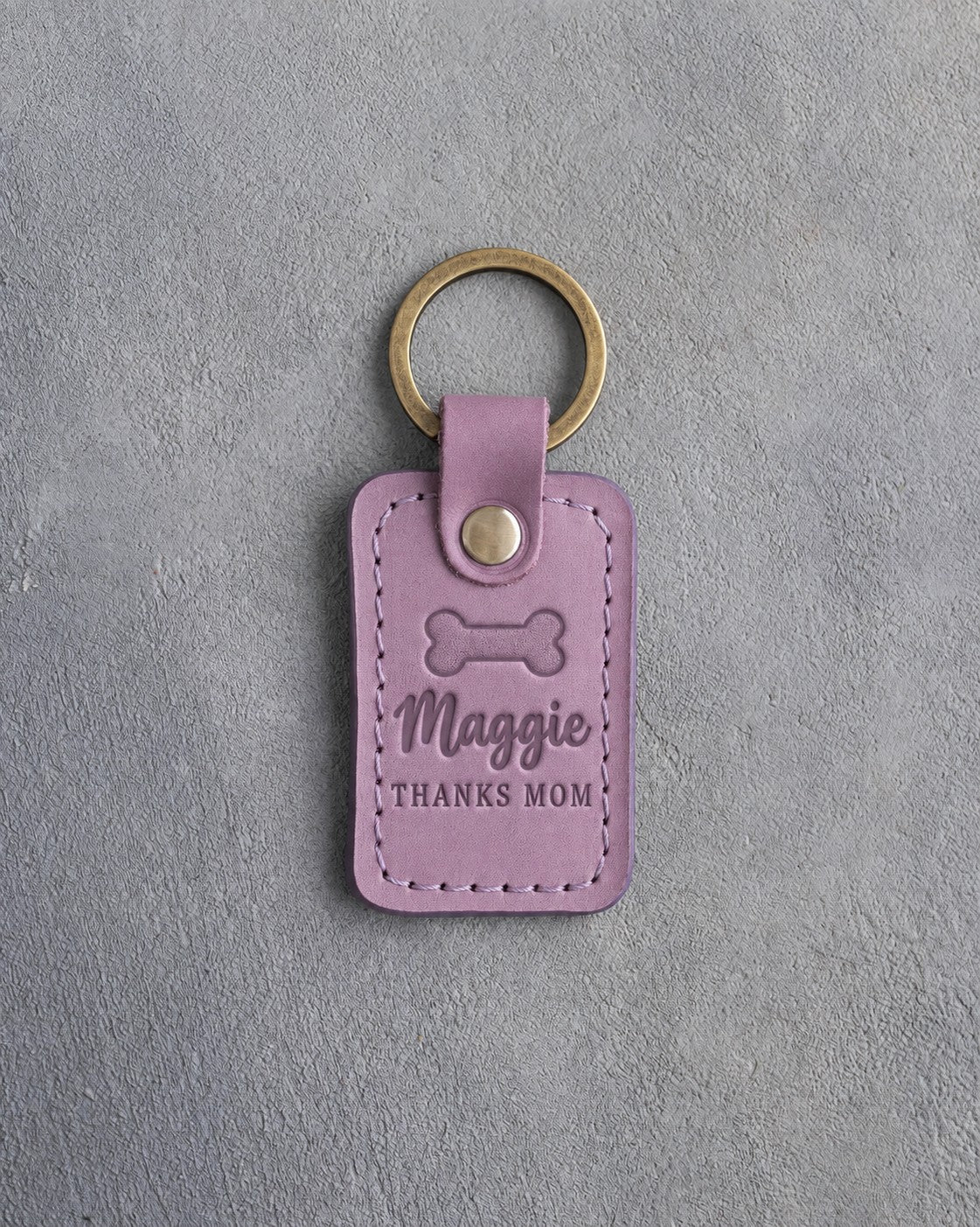 Lilac leather keychain debossed with a bone biscuit above Maggie and THANKS MOM, on gray concrete