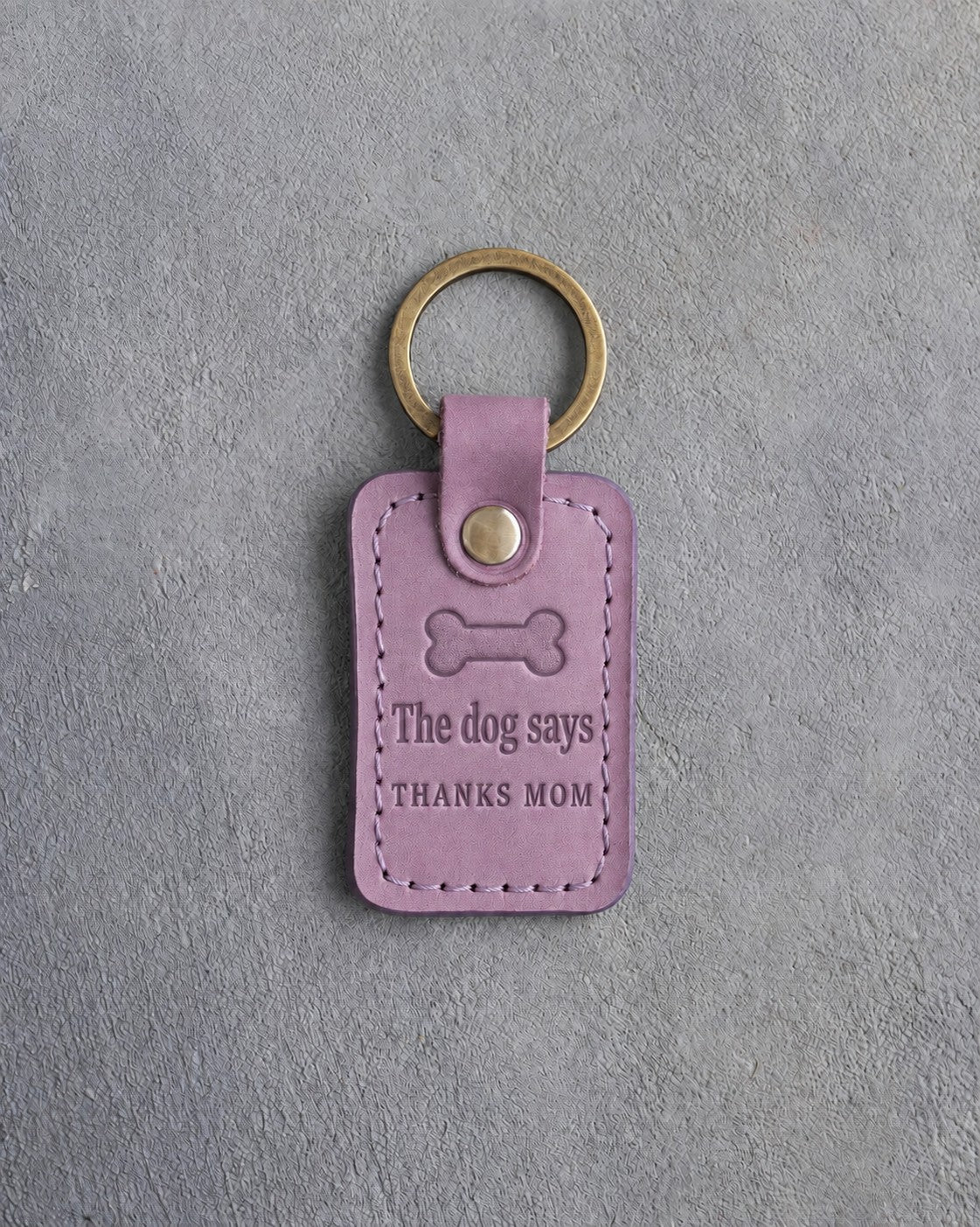 Lilac leather keychain debossed with a bone biscuit above The dog says and THANKS MOM, on gray concrete