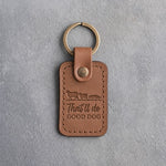 Caramel leather keychain debossed with two sheep and a crouching herding dog above the words That'll do GOOD DOG, on gray concrete
