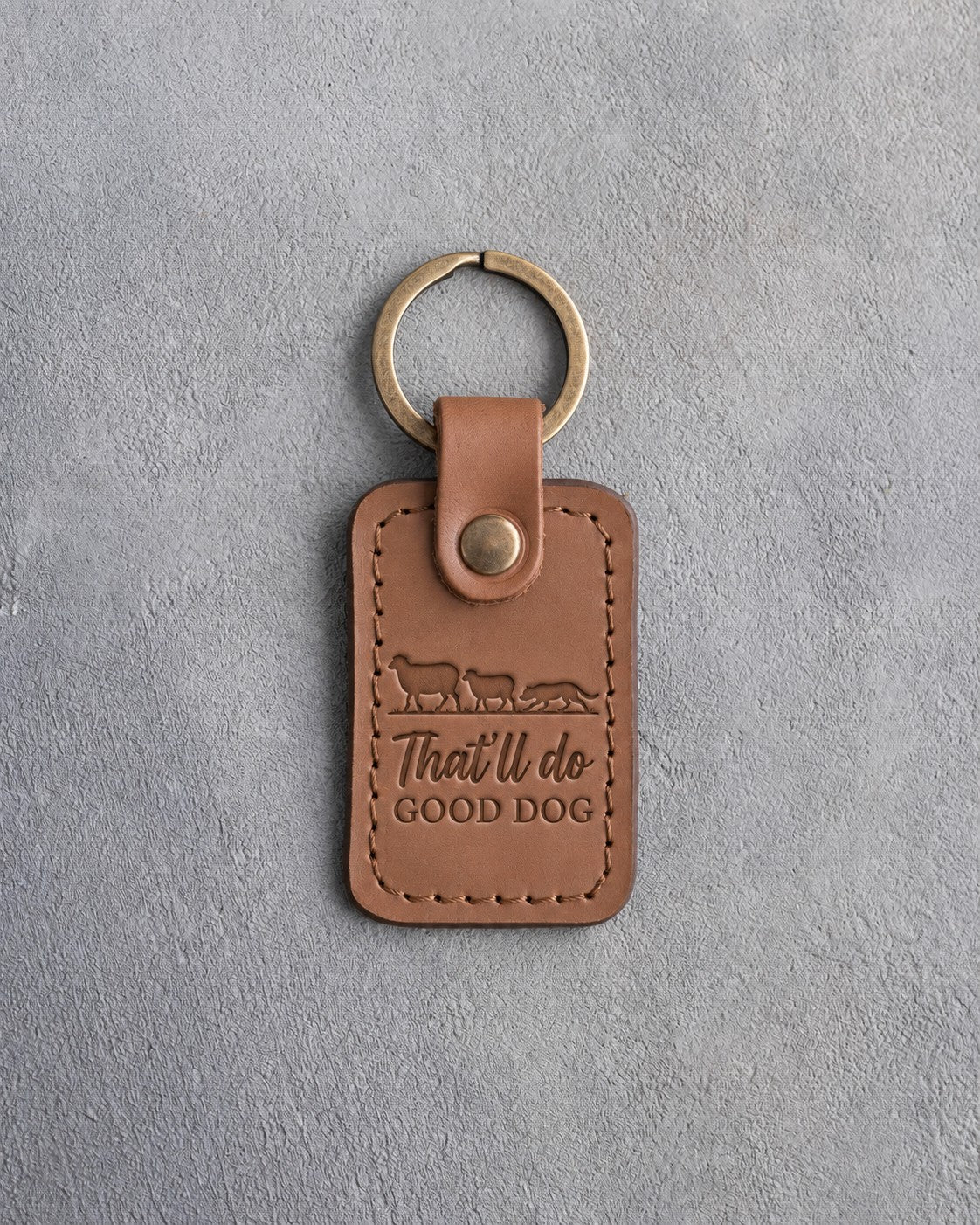 Caramel leather keychain debossed with two sheep and a crouching herding dog above the words That'll do GOOD DOG, on gray concrete