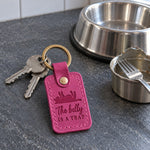 Fuchsia The Belly Is a Trap keychain on a soft rug beside a feather cat toy