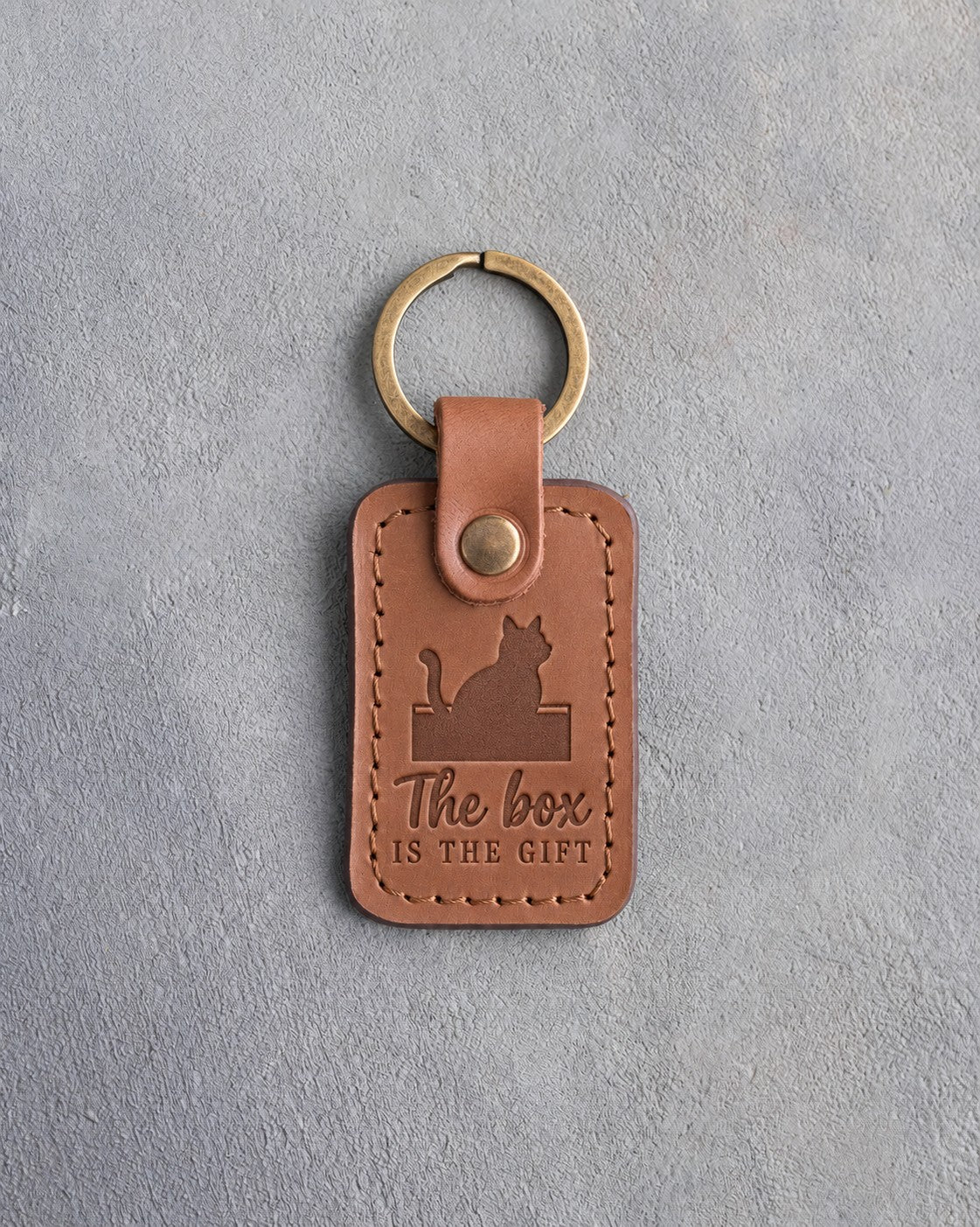 Caramel leather keychain debossed with a cat sitting in an open cardboard box above the words The box IS THE GIFT