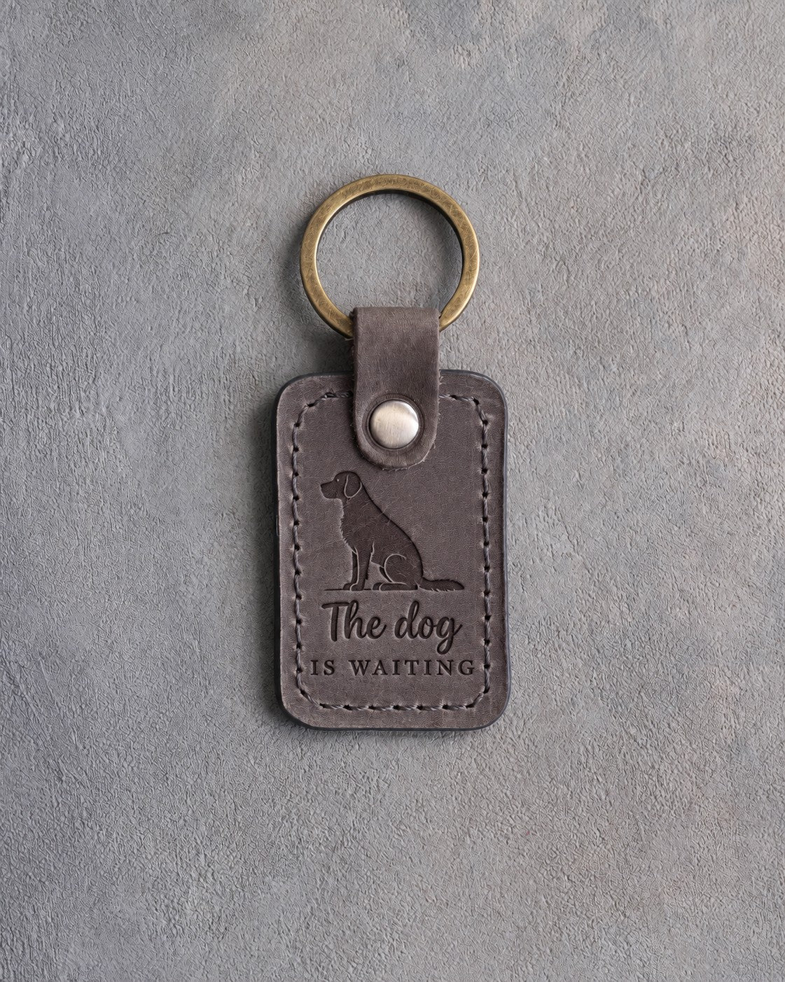 Country gray leather keychain debossed with a sitting mixed-breed dog and the words The dog IS WAITING, on gray concrete