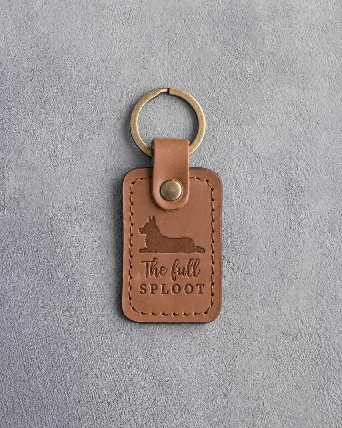 Caramel leather keychain debossed with a corgi lying flat in a sploot and the words The full SPLOOT, on gray concrete