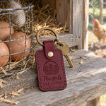 Wine red The Girls Are Laying keychain on a weathered pine nesting box ledge beside brown eggs in a wire basket, a hen soft in the run behind