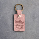 Blush pink leather keychain debossed with a dog resting its chin on a bedside and the words Therapy TEAM, on gray concrete