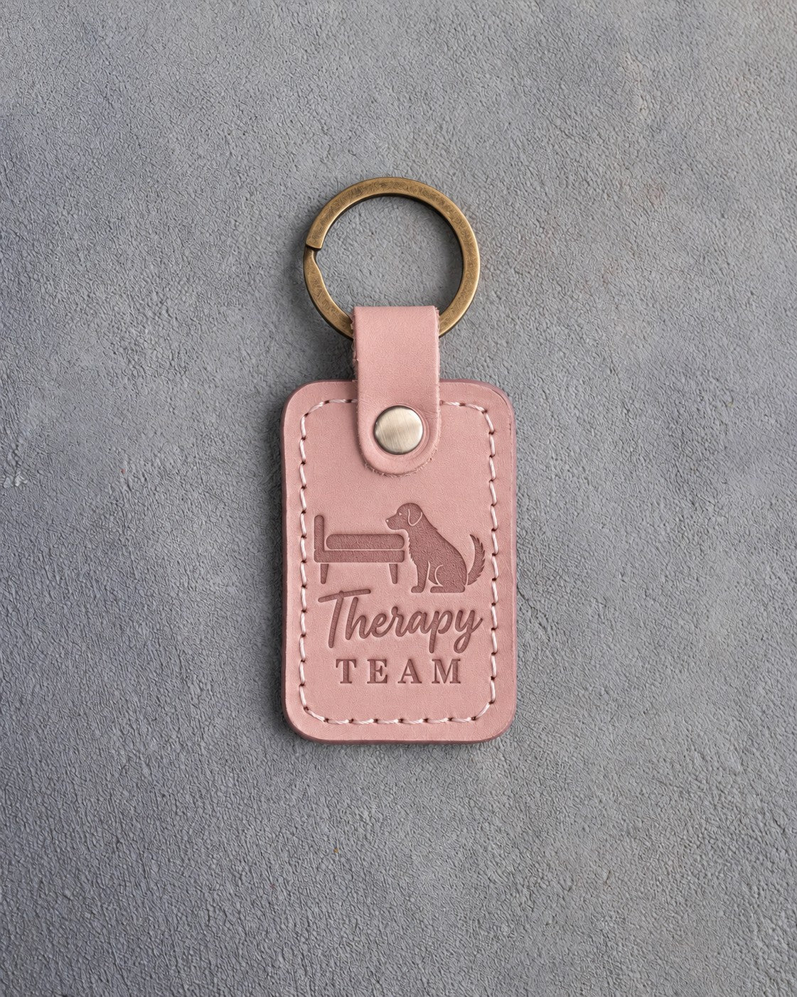 Blush pink leather keychain debossed with a dog resting its chin on a bedside and the words Therapy TEAM, on gray concrete