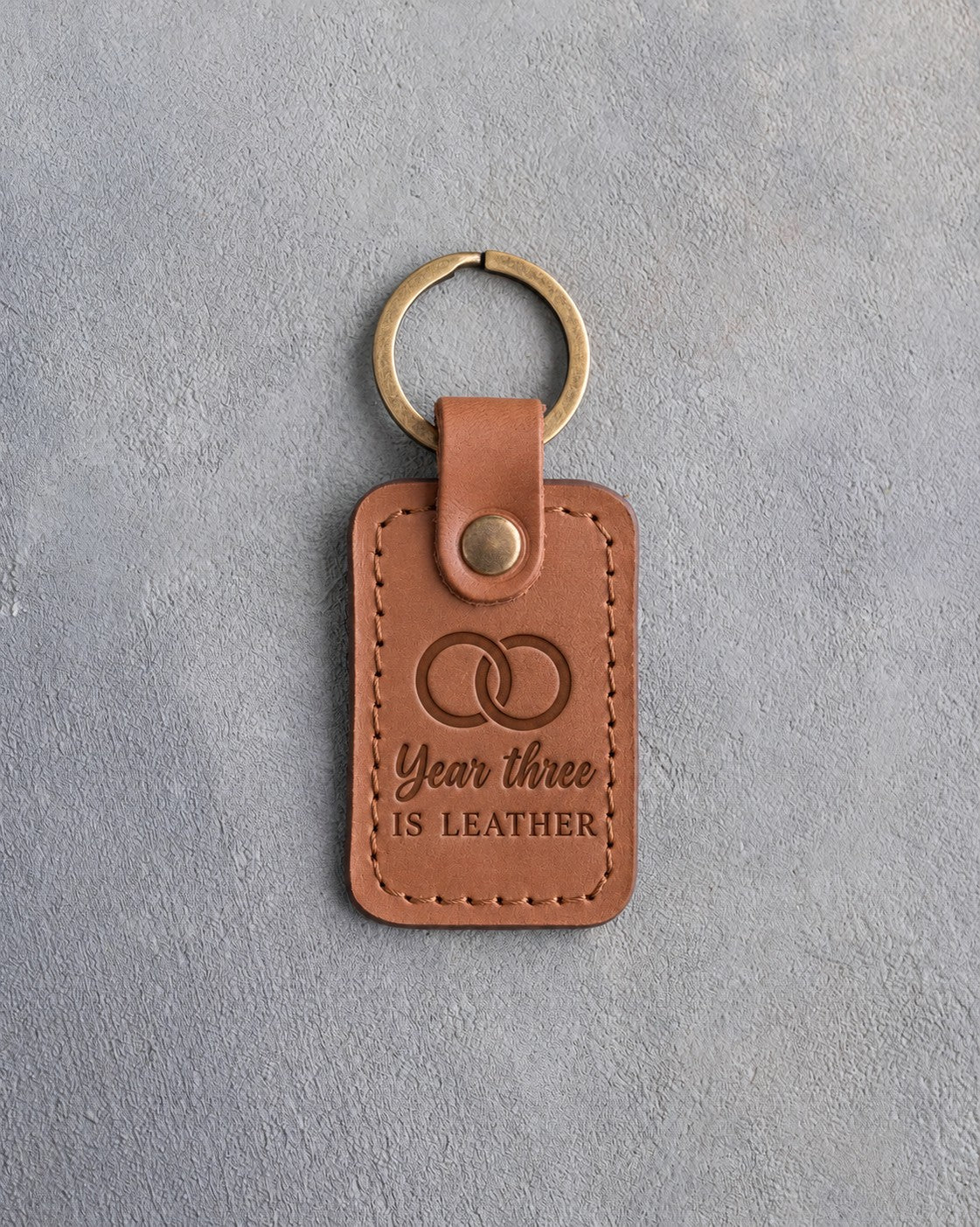 Caramel leather keychain debossed with two interlocked wedding rings above the words Year three IS LEATHER, on gray concrete