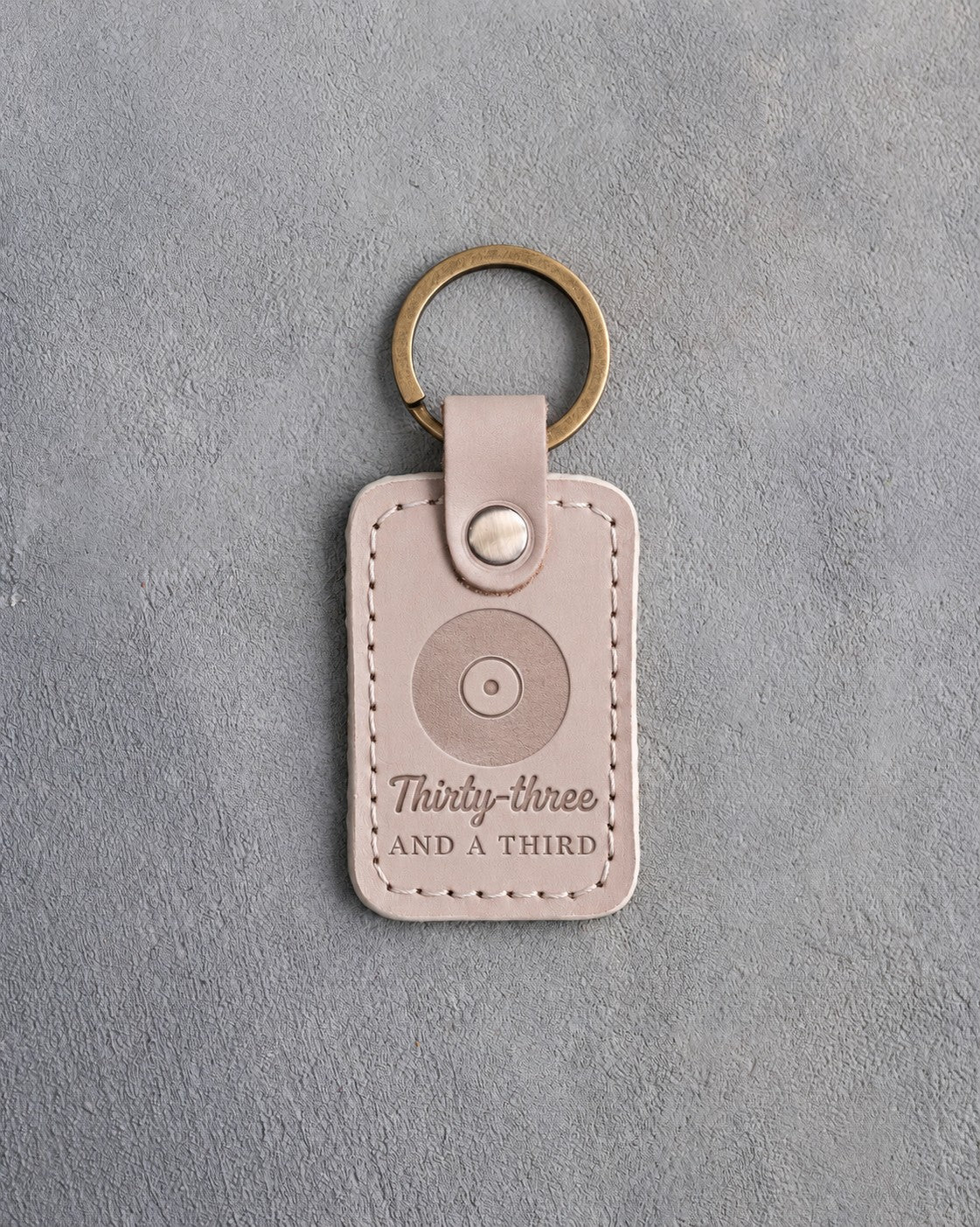 Gray sand leather keychain debossed with a vinyl record above the words Thirty-three AND A THIRD, on gray plaster