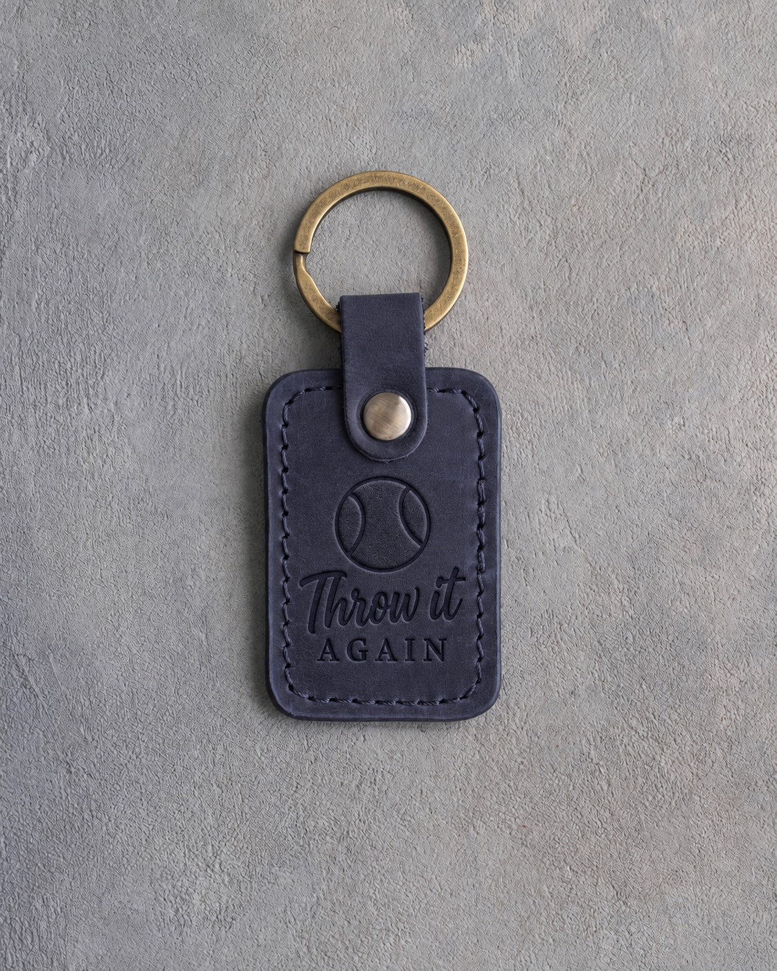 Blueberry navy leather keychain debossed with a tennis ball and the words Throw it AGAIN, on gray concrete