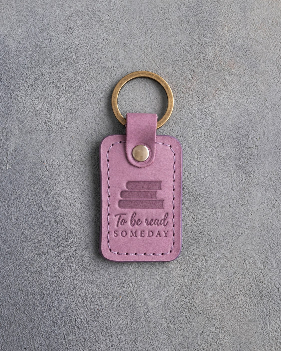 Lilac leather keychain debossed with a pile of three books above To be read and SOMEDAY, on gray concrete