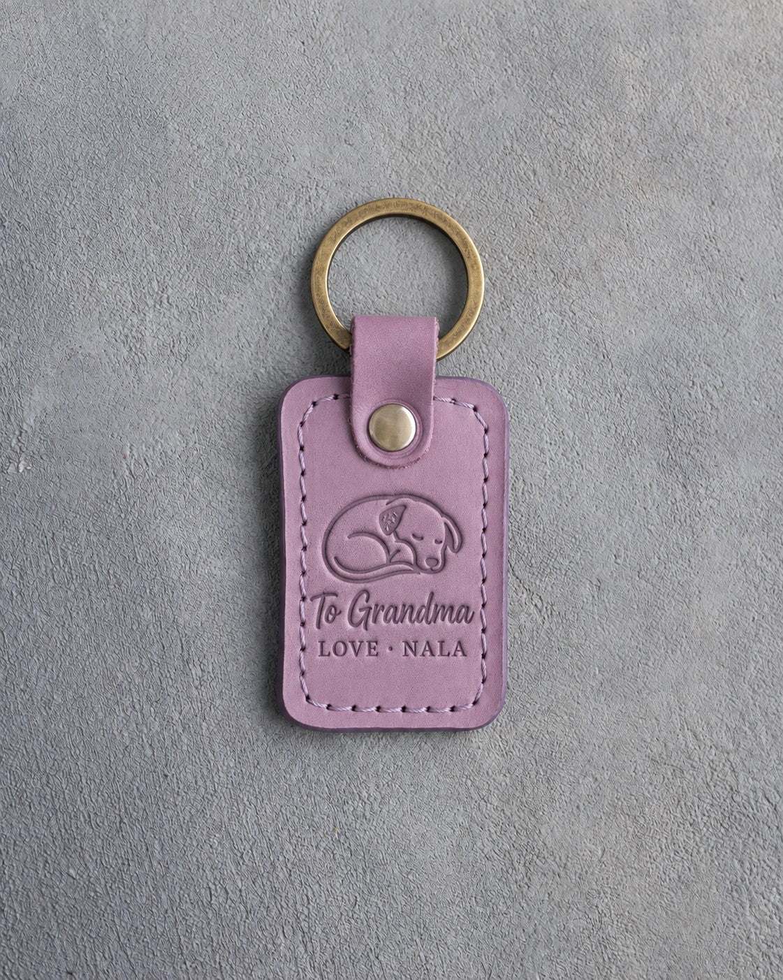 Lilac leather keychain debossed with a curled sleeping dog above To Grandma and LOVE · NALA, on gray concrete